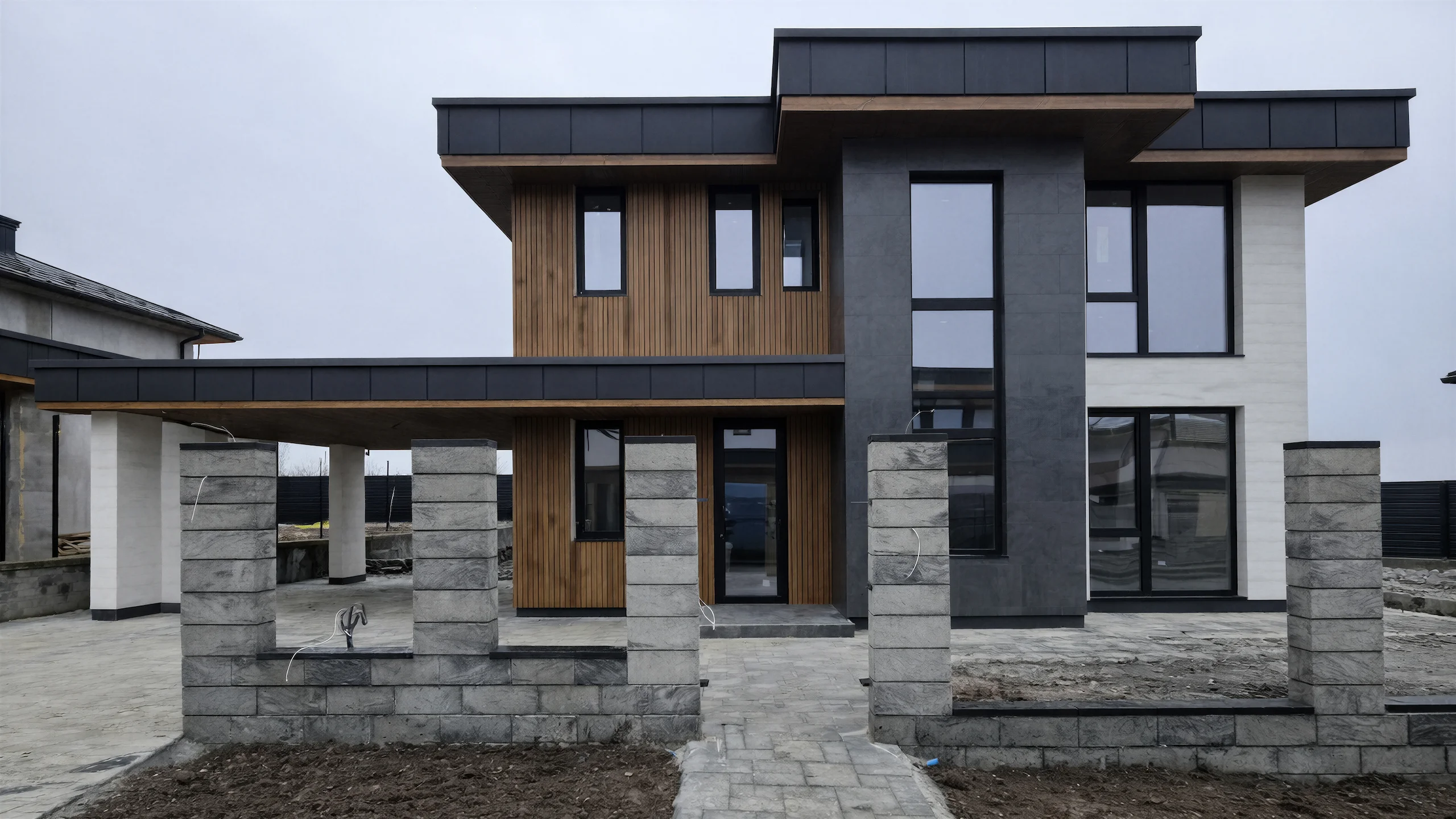 Главный фасад двухэтажного дома 240 м² с высоким темным объемом, деревянной входной зоной и каменной облицовкой