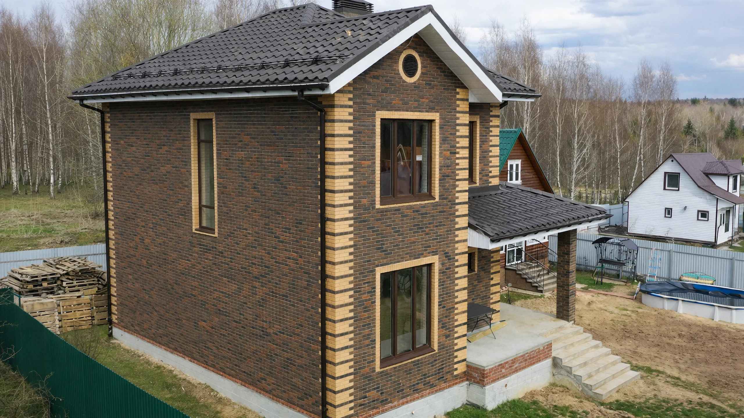 Боковой фасад двухэтажного дома 132 м² с темной кирпичной кладкой, высоким окном и вальмовой кровлей