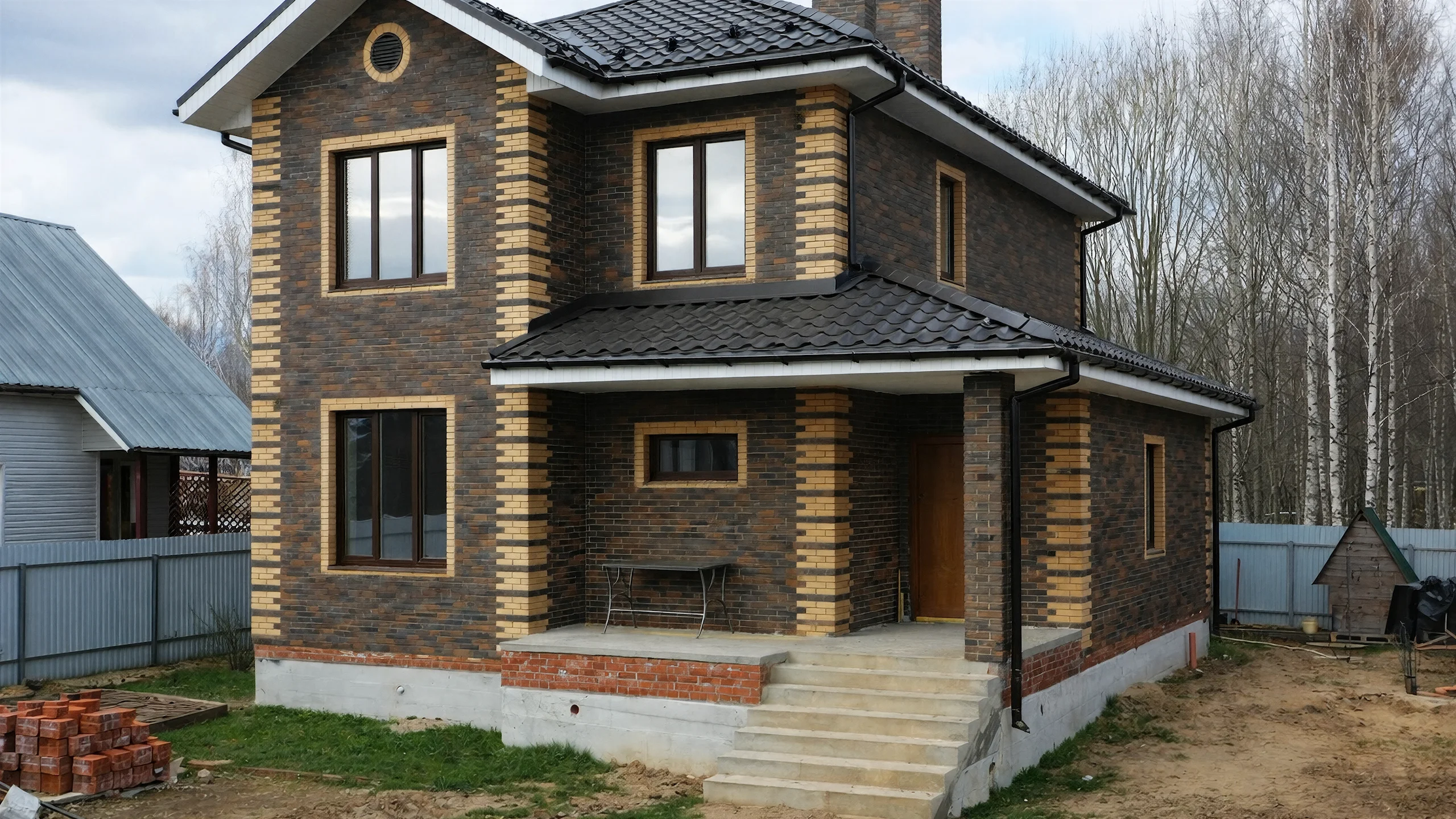 Главный фасад двухэтажного дома 132 м² с крытым входом, декоративными светлыми углами и темной кровлей
